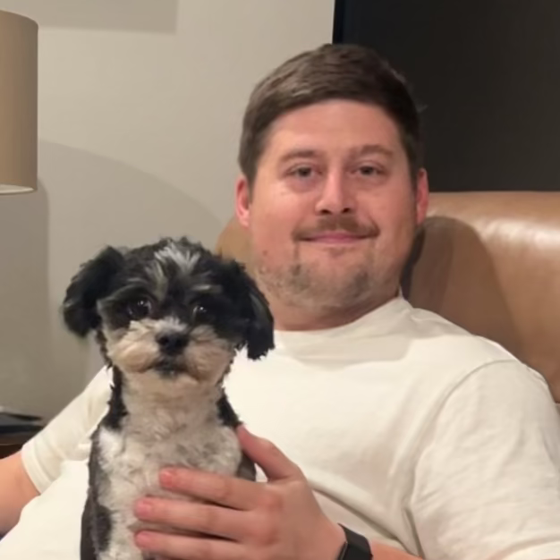 Man sitting on a brown recliner holding a small dog in a living room.